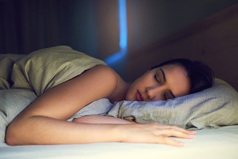 3 Health Benefits for Using Your AC While Sleeping. Photo of a young woman fast asleep in her bed.