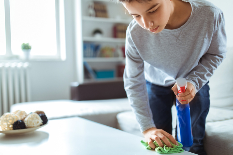 How to Reduce Dust in Your Home. Photo of a young boy wiping off a table with a cloth in his living room.